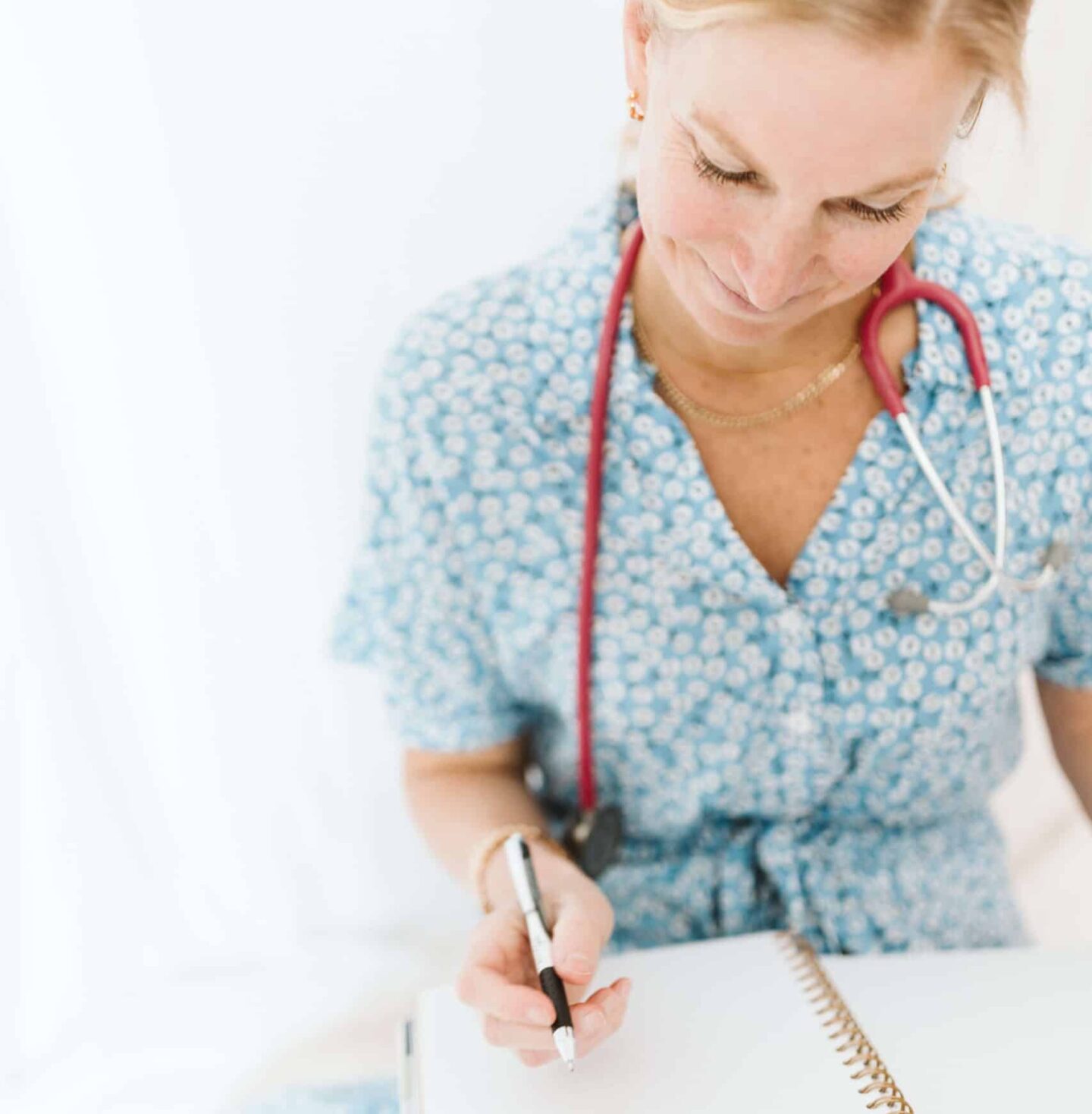 Image of Dr. Joyce sitting in a white airy room, wearing a blue sundress and her stethoscope, taking notes.