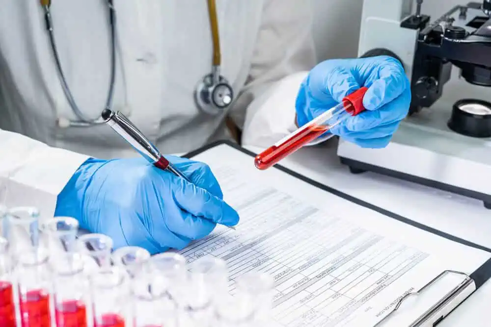 Close crop image of a lab tech in blue gloves holding a vial of blood, taking notes