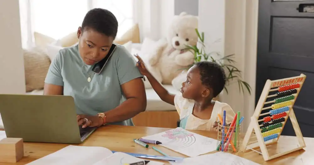 A busy parent trying to work on her laptop, while on the phone, with their young child asking for attention.